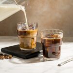 Cold brew vs iced latte side by side in glass cups with milk being poured over ice on a marble surface