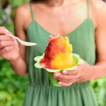 Hawaiian shave ice vs snow cone showing a woman holding a colorful strawberry and mango flavored shave ice in a green cup outdoors