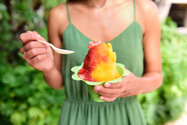 Hawaiian shave ice vs snow cone showing a woman holding a colorful strawberry and mango flavored shave ice in a green cup outdoors