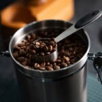 Coffee beans in an opaque, airtight stainless steel canister with a scoop, showing how to store coffee beans properly
